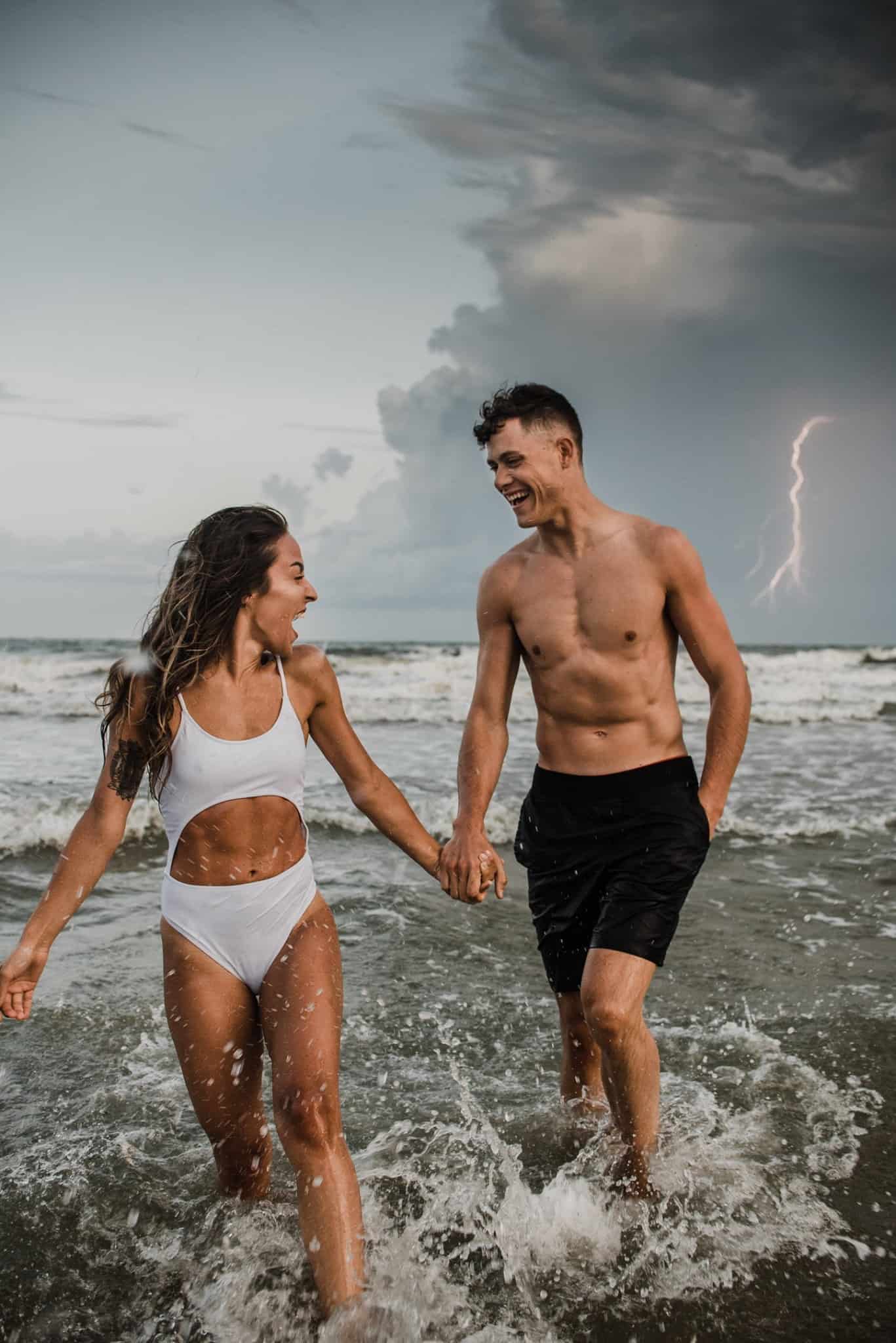 Man and woman holding hands in splashing seas water, looking at each other smiling. Lighting strike in background