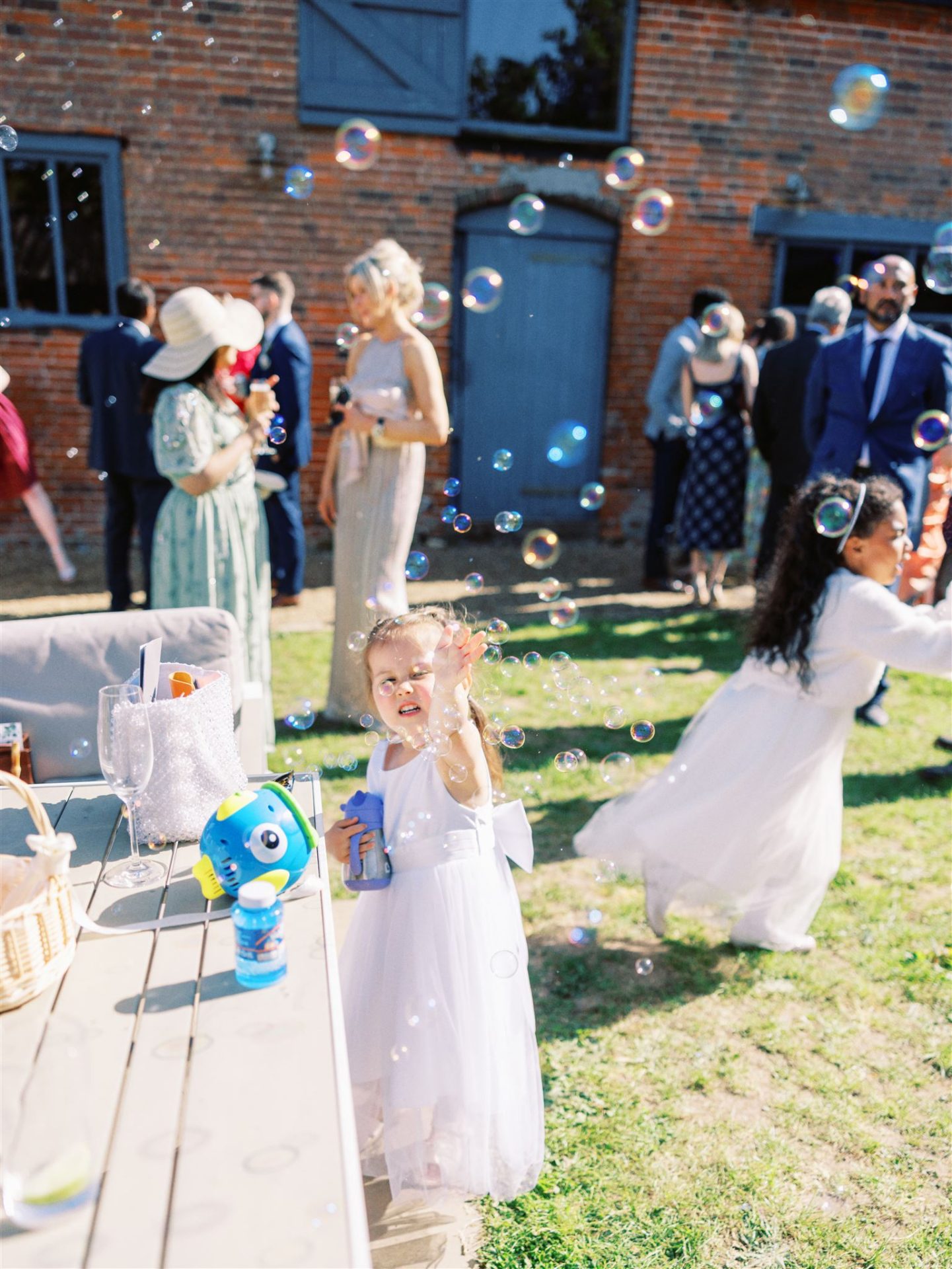 Little girls in white bridesmaid dresses playing in front of a bubble machine