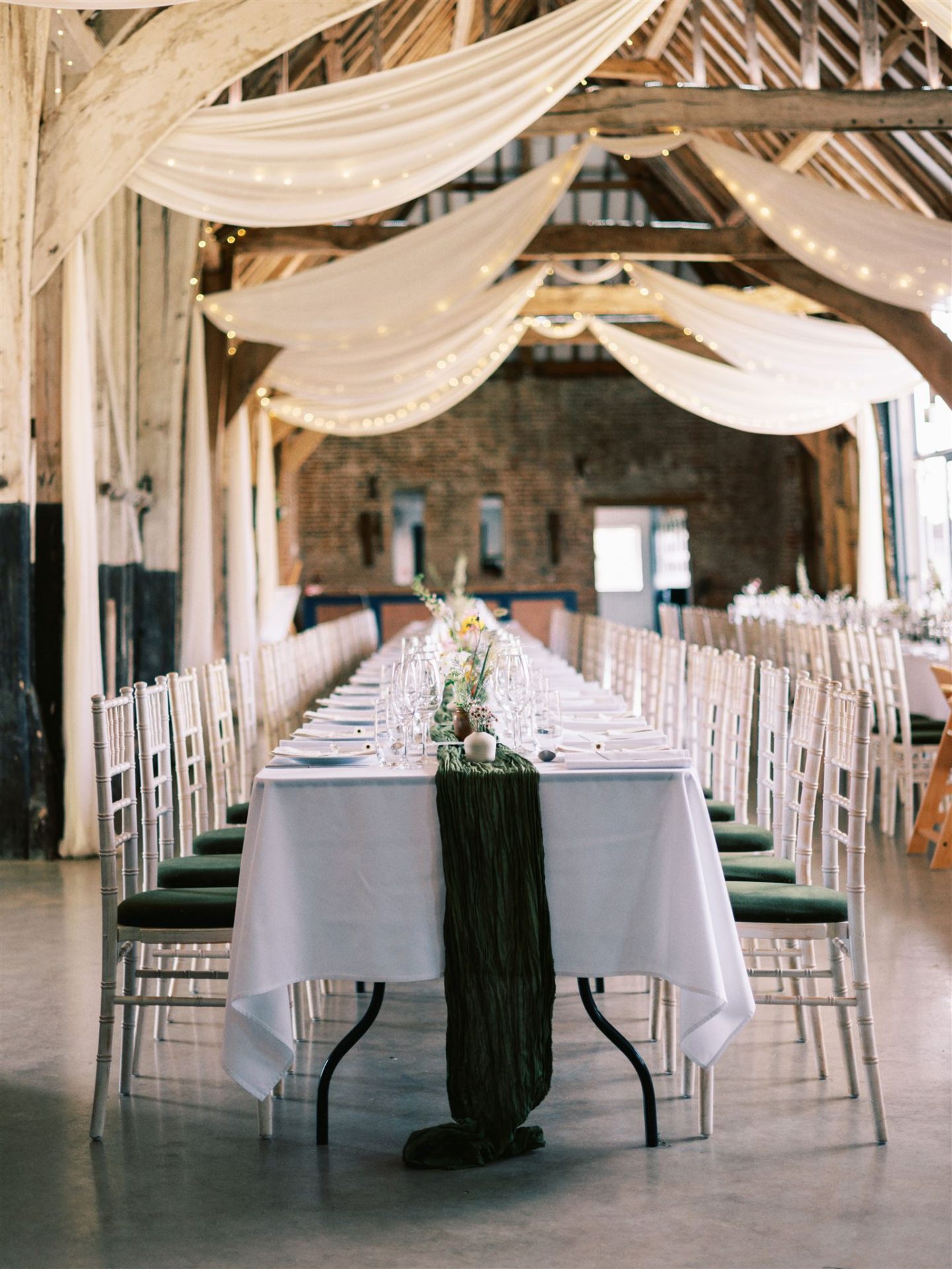 rustic barn venue with ceiling drapery and banqueting table with white linen, forest green runner and small yellow and pink bud vases