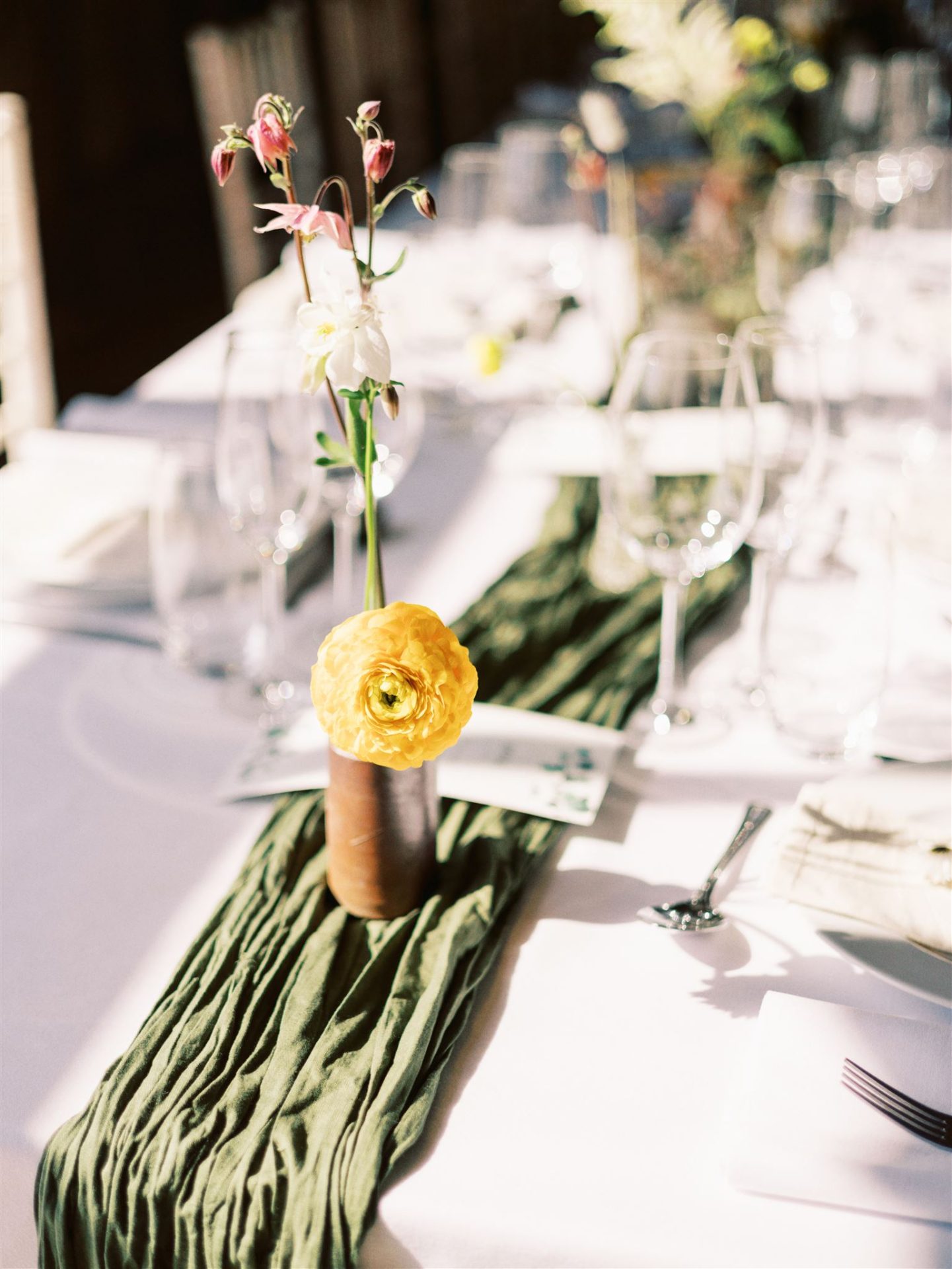 white linen, forest green table runner and stone bud vases with small yellow flower in it