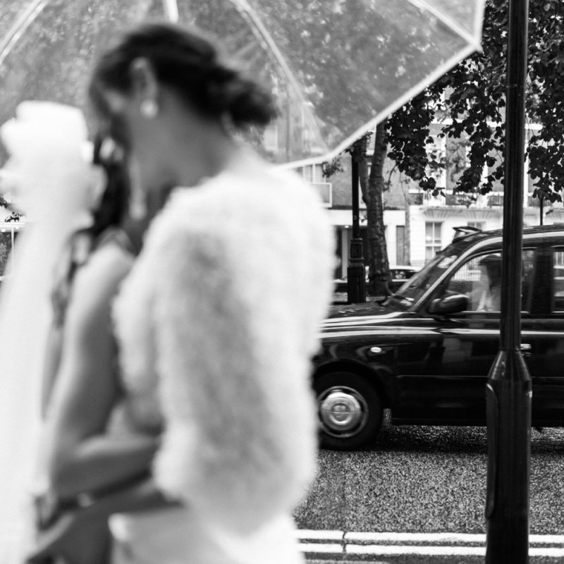 Blurred image, two brides hugging under umbrella. Black and white cab driving in the background